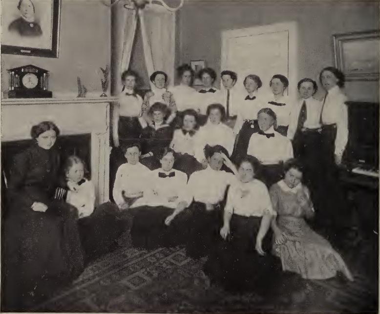 Black and white photograph showing 20 people. All three rows. Most are wearing white tops and dark bottoms. A woman in a dark dress sits to the right side of the photo in front of a fireplace.