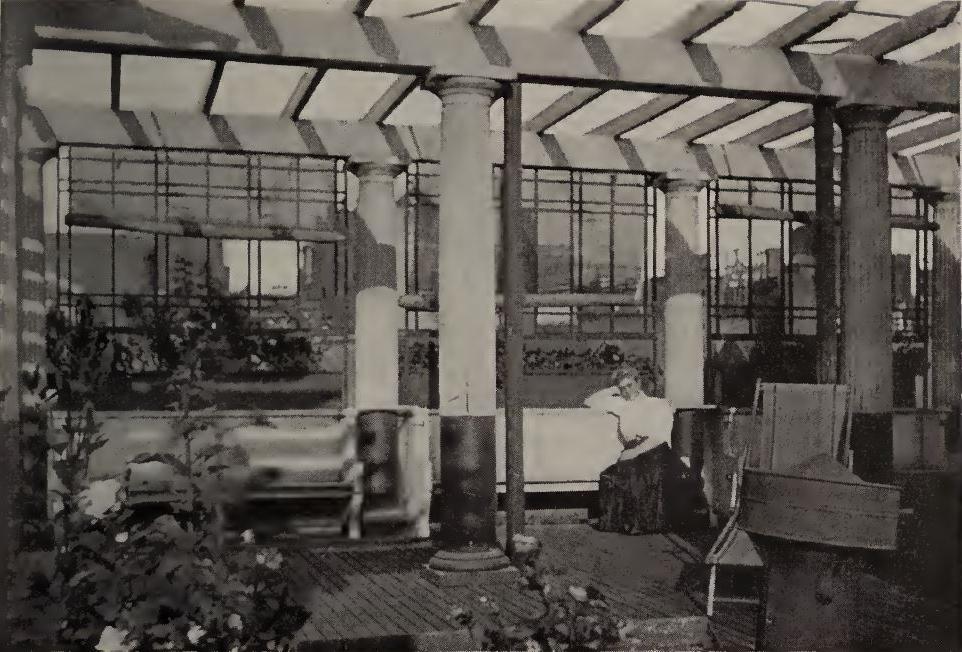 black and white photo taken from within the rooftop garden at 44 Chambers Street. A woman dressed in a white shirt and long, dark skirt sits on a bench in the photo. There are 4 full columns visible holding horizontal beams (a pergola). Several plants are also visible. 